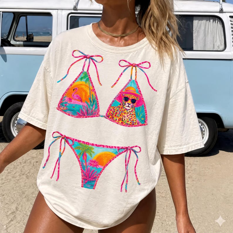 May include: Cream-colored t-shirt featuring a colorful bikini design. The bikini top showcases a flamingo and a leopard wearing sunglasses, while the bottom displays a tropical sunset. The design incorporates pink, blue, and orange hues.