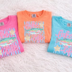 May include: Three colorful t-shirts: pink, orange, and blue. Each shirt features a design with a rainbow trout, the words "Lake Life" in pink script, blue stripes, and gold star accents. The shirts are folded on a white, fluffy surface.