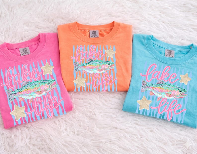 May include: Three colorful t-shirts: pink, orange, and blue. Each shirt features a design with a rainbow trout, the words "Lake Life" in pink script, blue stripes, and gold star accents. The shirts are folded on a white, fluffy surface.