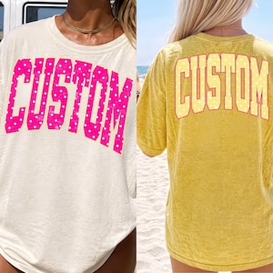 May include: Two oversized t-shirts, one cream and one yellow, each with the word "CUSTOM" in a bold, block letter font. The cream shirt has pink polka dots within the letters, while the yellow shirt has yellow polka dots.