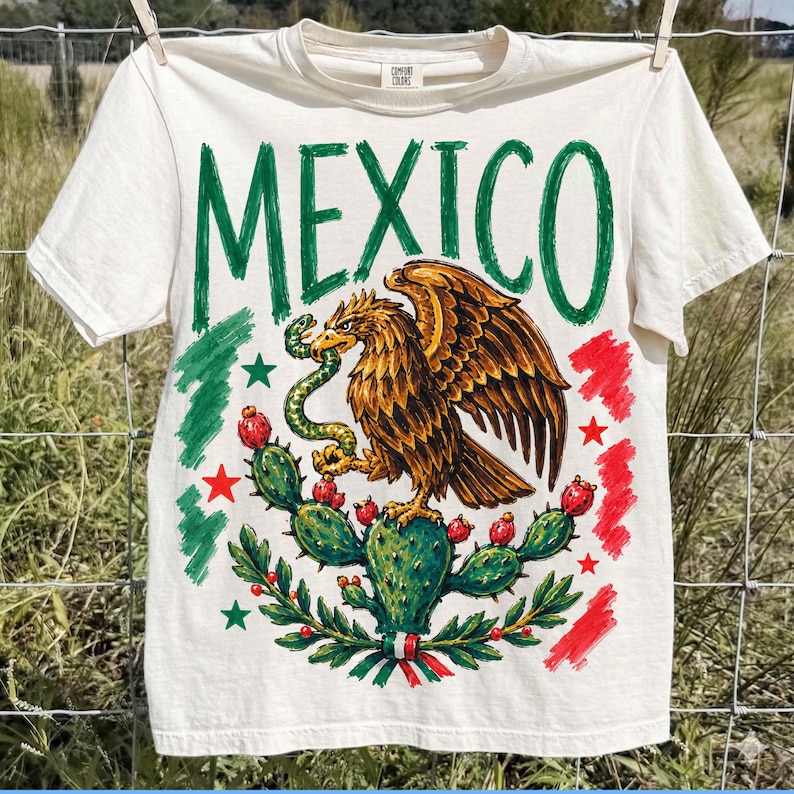 May include: A cream-colored short-sleeve t-shirt featuring the word MEXICO in large green letters. The graphic depicts a golden eagle perched on a cactus, holding a green snake in its beak, surrounded by red and green brushstroke accents and stars.