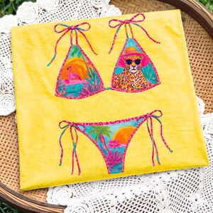 May include: A yellow bikini set with a tropical design. The top features a flamingo and a leopard wearing sunglasses. The bottom has a sunset and palm trees. The bikini has pink and blue accents.
