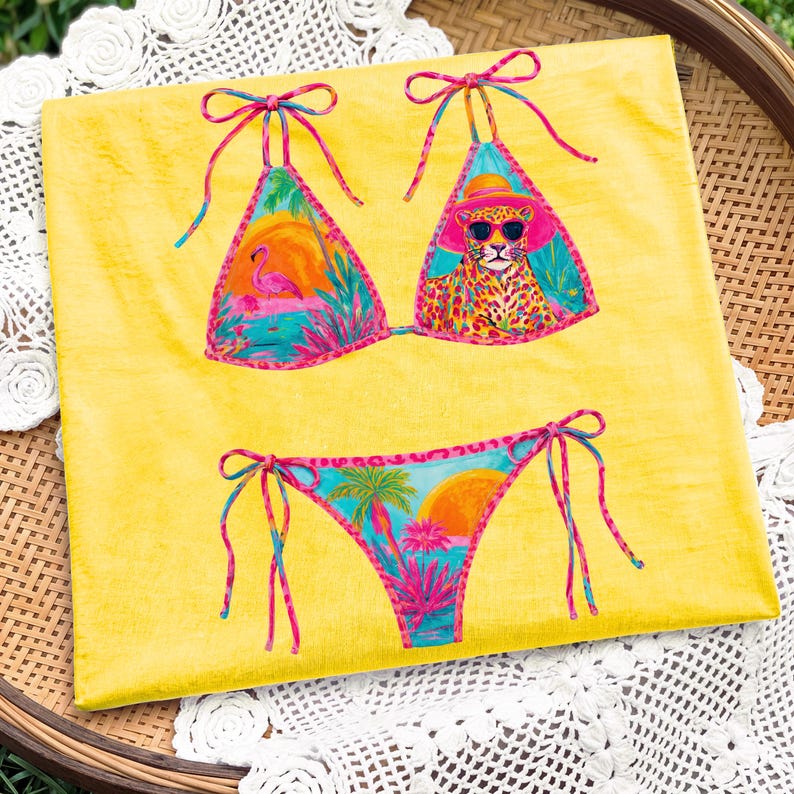 May include: A yellow bikini set with a tropical design. The top features a flamingo and a leopard wearing sunglasses. The bottom has a sunset and palm trees. The bikini has pink and blue accents.