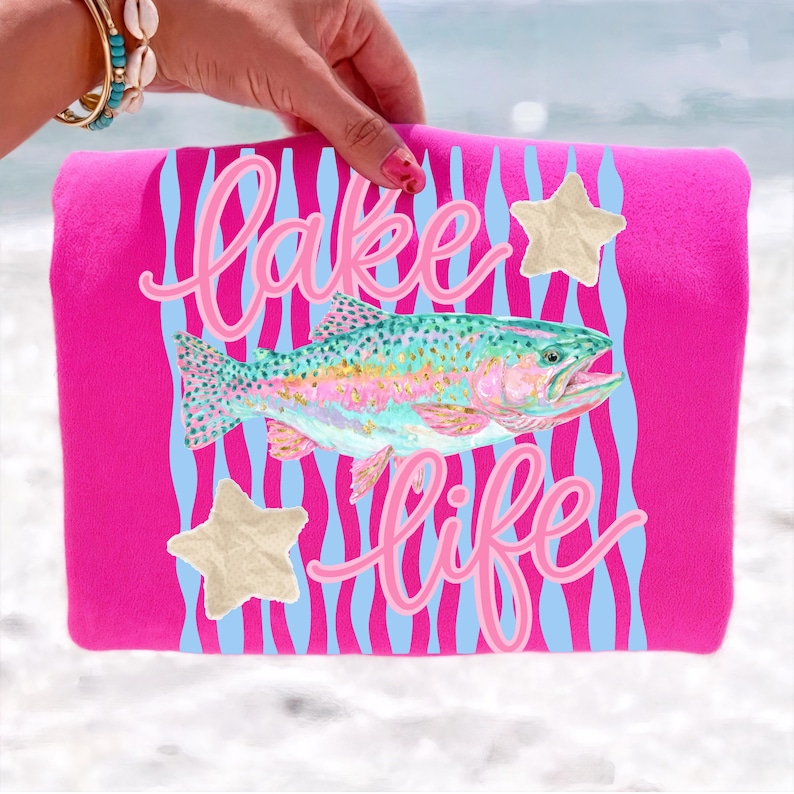 May include: A hot pink beach towel with the words "Lake Life" in cursive script and a colorful fish illustration. The towel also features light blue wavy stripes and two beige star accents.
