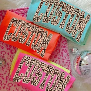 May include: Four colorful t-shirts in shades of orange, pink, teal, and yellow. Each shirt features the word "CUSTOM" in a leopard print design. The shirts are arranged on a pink and white patterned surface, with disco balls.