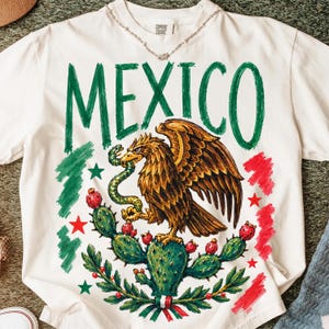 May include: A white short-sleeve t-shirt featuring a large graphic of an eagle perched on a cactus holding a snake. The word MEXICO is printed in large green letters above the eagle. The design includes red and green brushstroke accents.