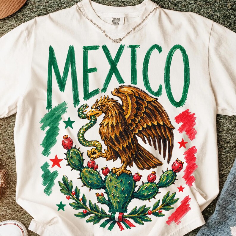 May include: A white short-sleeve t-shirt featuring a large graphic of an eagle perched on a cactus holding a snake. The word MEXICO is printed in large green letters above the eagle. The design includes red and green brushstroke accents.