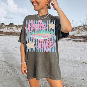 May include: Dark olive green t-shirt with a colorful fish graphic and the words "Lake Life" in pink script. The design includes blue stripes and star accents. The shirt is a casual, oversized fit.