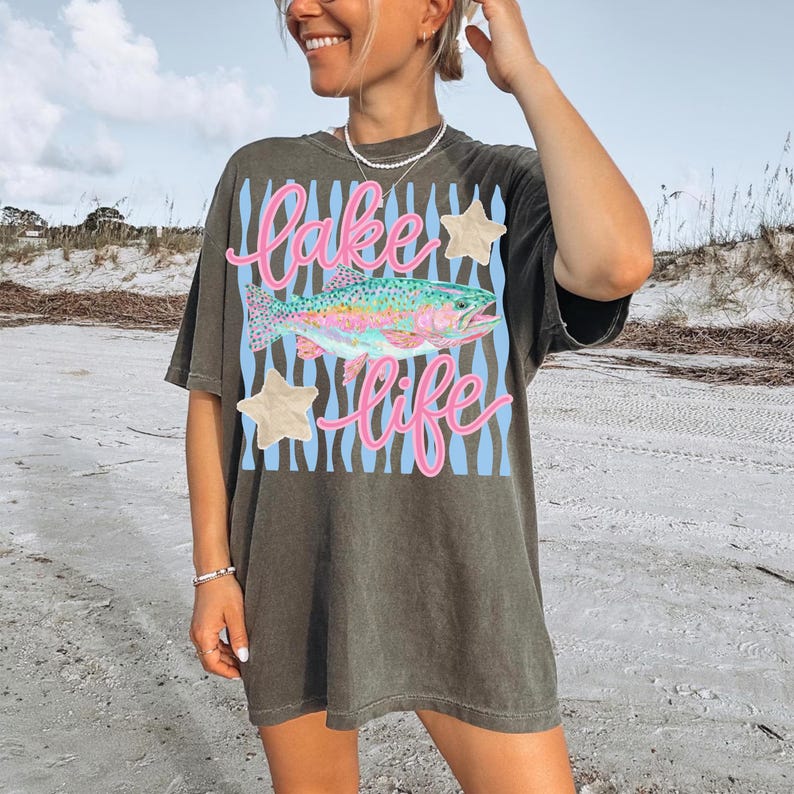 May include: Dark olive green t-shirt with a colorful fish graphic and the words "Lake Life" in pink script. The design includes blue stripes and star accents. The shirt is a casual, oversized fit.