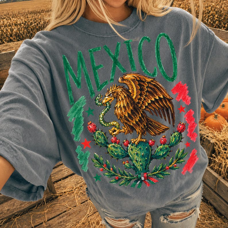 May include: A slate gray short-sleeve t-shirt featuring a graphic print of the word MEXICO in green letters above an illustration of a golden eagle perched on a cactus while holding a green snake, accented with red and green brushstroke details.