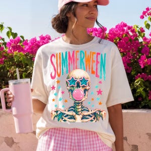 May include: Cream-colored t-shirt with the word "SUMMERWEEN" in colorful letters. The shirt features a skeleton wearing star-shaped sunglasses and blowing a bubblegum bubble. Pink and blue stars surround the skeleton.