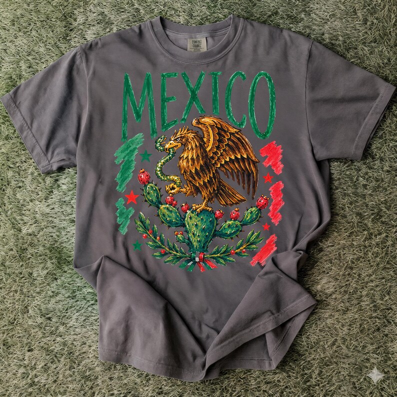 May include: A charcoal gray short-sleeve t-shirt featuring a graphic print of a golden eagle perched on a cactus while holding a snake. The word MEXICO is printed in large green letters above the eagle, flanked by red and green brushstroke accents.