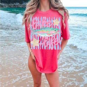 May include: A coral-colored t-shirt with the words "Lake Life" in a cursive font, a rainbow trout, and two gold star accents. The design is set against a light blue striped background.