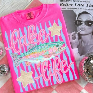 May include: A hot pink t-shirt with a graphic design featuring the words "Lake Life" in pink script, a colorful fish, and two beige star-shaped patches. The background has blue and pink stripes.