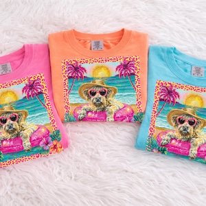 Puede incluir: Tres camisetas coloridas: rosa, naranja y azul. Cada camiseta presenta un gráfico de un perro con gafas de sol y un sombrero de paja, descansando en un flotador inflable rosa en la playa. El gráfico incluye palmeras, una puesta de sol y un borde con estampado de leopardo.