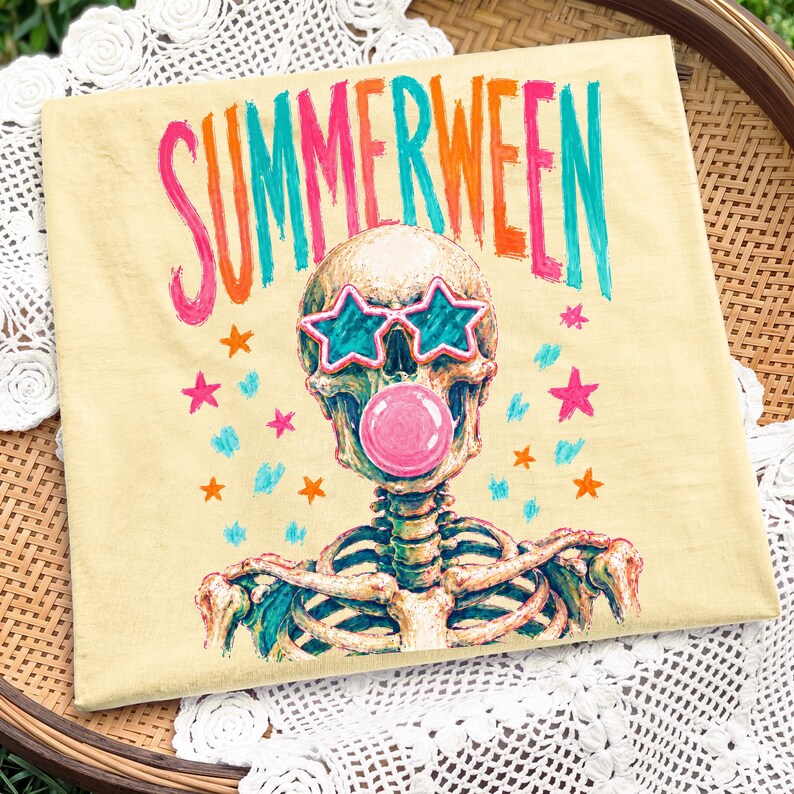 May include: A light yellow t-shirt with the word "SUMMERWEEN" in pink, orange, and teal lettering. The shirt features a skeleton wearing star-shaped sunglasses and blowing a pink bubble. Colorful stars are scattered around the design.