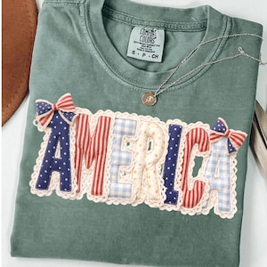 Op de afbeelding: Een saliegroen t-shirt met het woord "AMERICA" op de voorkant. De letters zijn in rood, wit en blauwe patronen met een kanten rand. Een zilveren ketting met een kleine hanger ligt op het shirt.