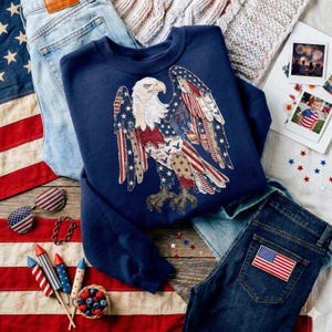 May include: A navy blue sweatshirt with an American flag eagle design, alongside blue jeans, sunglasses, and patriotic decorations. The image includes a small basket of berries and fireworks.