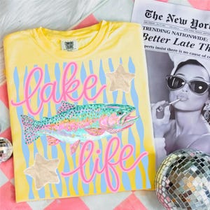 May include: Yellow t-shirt with a colorful fish graphic and the words "Lake Life" in pink script. The design includes blue vertical stripes and star accents. A newspaper and disco ball are in the background.