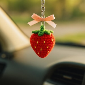 May include: A handmade, red crocheted strawberry car charm with a green stem and yellow seeds. The charm hangs from a silver chain and is adorned with a small, light pink bow. The charm is hanging from a car's rearview mirror.