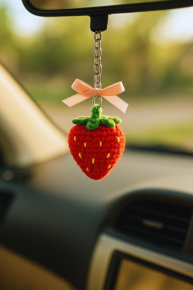 May include: A handmade, red crocheted strawberry car charm with a green stem and yellow seeds. The charm hangs from a silver chain and is adorned with a small, light pink bow. The charm is hanging from a car's rearview mirror.