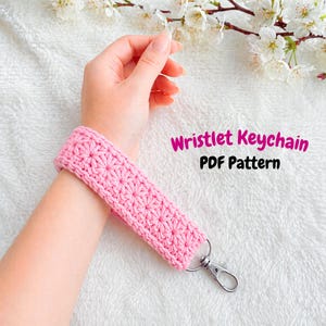 Puede incluir: Un llavero de pulsera de ganchillo rosa con un diseño de estrella. La pulsera está unida a un cierre plateado. El texto en la imagen dice "Wristlet Keychain PDF Pattern".