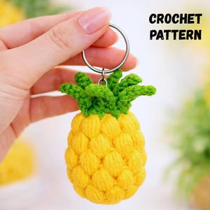 Pode incluir: Um chaveiro de abacaxi de crochê amarelo e verde feito à mão. O abacaxi tem uma superfície texturizada e um topo verde e frondoso. O chaveiro é preso a um anel de metal prateado. O texto na imagem diz "CROCHET PATTERN".