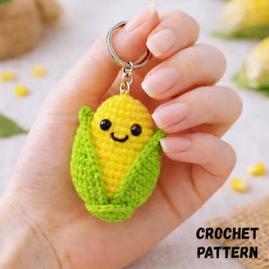 Puede incluir: Un llavero de maíz tejido a crochet con una cara sonriente, con maíz amarillo y cáscara verde. El llavero está sujeto a un anillo plateado. El texto "CROCHET PATTERN" está en la esquina inferior derecha.