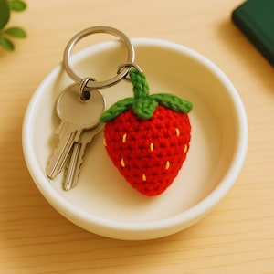May include: A red crocheted strawberry keychain with a green stem and leaves, attached to a silver keyring with two keys. The keychain is in a white bowl.