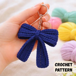 May include: A navy blue crochet bow keyring with a silver ring. The bow is handmade with a textured stitch. The background includes colourful yarn skeins. The text "CROCHET PATTERN" is visible.