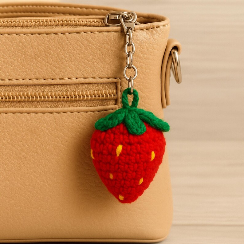 May include: A red crocheted strawberry keychain with green leaves and yellow seeds hangs from a silver chain attached to a tan purse. The strawberry is a vibrant, handmade accessory.