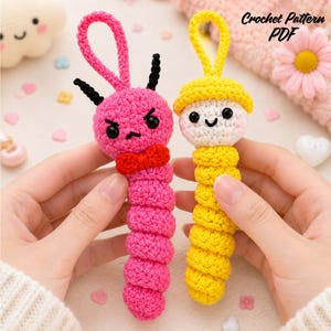 May include: Two crochet worm keychains with hanging loops. The pink worm has black antennae, a red bow tie, and an angry expression. The yellow worm wears a yellow hat and has a white face with a smiling expression. Text reads Crochet Pattern PDF.