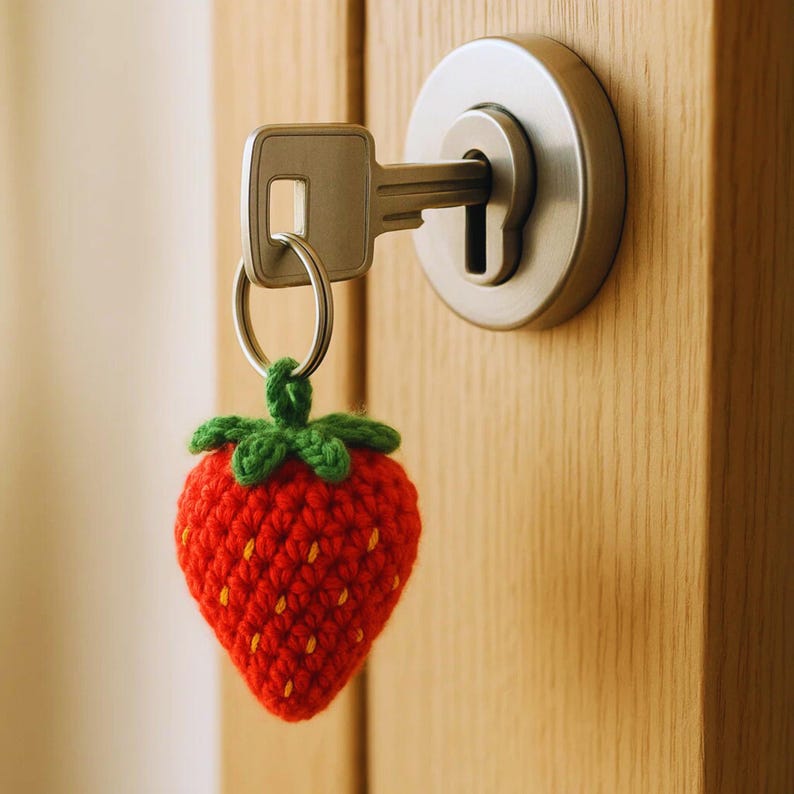 May include: A red crocheted strawberry keychain with green leaves and yellow seeds hangs from a silver key. The key is inserted into a silver doorknob on a wooden door.