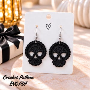 Puede incluir: Un par de pendientes de calavera tejidos a ganchillo en color negro, sujetos a una tarjeta de presentación blanca con un pequeño icono de corazón negro. Los pendientes cuelgan de ganchos de metal plateado. El texto "Crochet Pattern ENG PDF" es visible.