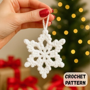 May include: A white, crocheted snowflake ornament with a loop for hanging. The snowflake is held up against a blurred background of a Christmas tree with lights and wrapped gifts. The text "CROCHET PATTERN" is in the lower right corner.