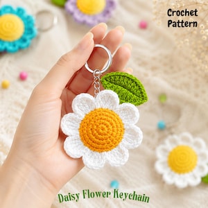May include: A crochet daisy flower keychain featuring a bright yellow center, white petals, and a green leaf attached to a silver metal ring. The text Crochet Pattern and Daisy Flower Keychain are visible in the image along with other flowers.