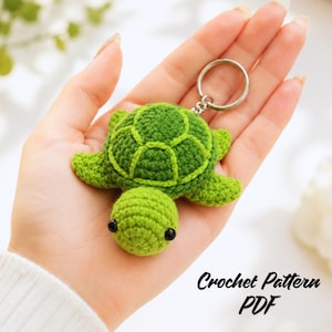 Puede incluir: Un pequeño llavero de tortuga hecho a mano en ganchillo con cabeza verde claro y caparazón verde oscuro con segmentos cosidos visibles. La tortuga tiene dos pequeños ojos de cuentas negras y está unida a una anilla metálica plateada. Texto Crochet Pattern PDF.