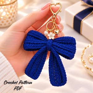 May include: A deep blue crocheted bow keyring featuring a gold-toned heart-shaped clasp, a small white flower charm with a central crystal, a single white pearl bead, and a small gold heart charm attached to a gold ring connector.