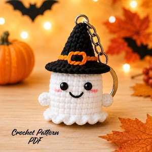 May include: A white crocheted ghost keychain wearing a black witch hat with an orange band and buckle. The ghost has black bead eyes, a stitched smile, and pink blush marks. A metal keyring is attached to the top of the hat. Text reads Crochet Pattern PDF.