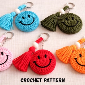 Op de afbeelding: Een verzameling handgemaakte gehaakte smiley-sleutelhangers in verschillende kleuren, waaronder blauw, groen, roze, rood en oranje. Elke sleutelhanger heeft een kwastje en een metalen ring. De tekst "CROCHET PATTERN" staat onderaan.