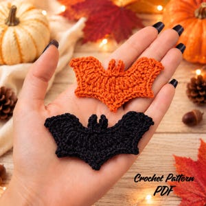May include: Two crocheted bat appliques, one in bright orange and one in solid black, featuring detailed wing textures and small ears. These flat, textile decorations are shaped like flying bats and are crafted with visible yarn stitching.