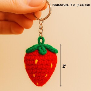 May include: A handmade red crochet strawberry keychain with green leaves and yellow seeds. The keychain is 2 inches (5 cm) tall and attached to a silver keyring. The text "Finished Size: 2 in (5 cm) tall" is visible.