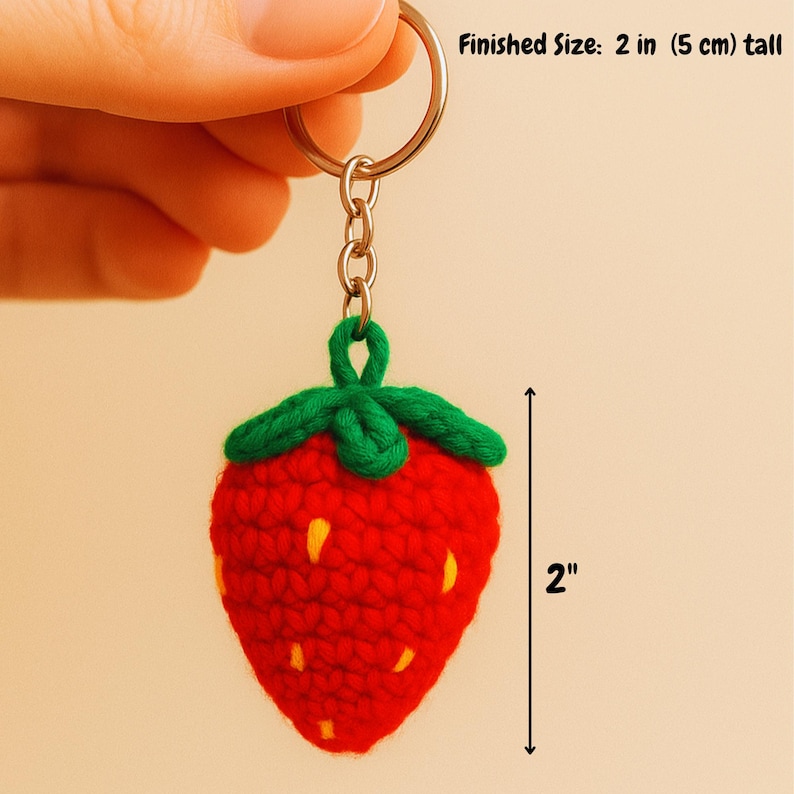May include: A handmade red crochet strawberry keychain with green leaves and yellow seeds. The keychain is 2 inches (5 cm) tall and attached to a silver keyring. The text "Finished Size: 2 in (5 cm) tall" is visible.