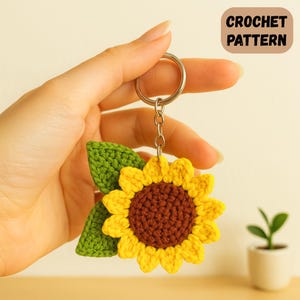 May include: A handmade crochet sunflower keychain with a yellow flower, brown center, and green leaves. The keychain has a silver ring and chain. The text "CROCHET PATTERN" is in a brown rectangle in the upper right corner.