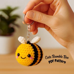 May include: A handmade yellow and black crochet bumble bee keychain with a smiling face and white wings. The keychain is held by a hand and has a silver keyring. The text "Cute Bumble Bee PDF Pattern" is visible.