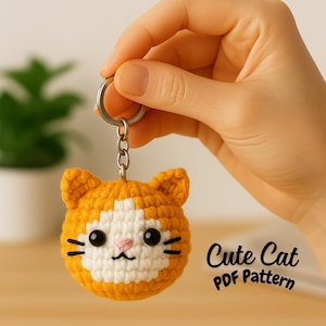 Op de afbeelding: Gehaakte oranje en witte katten sleutelhanger met zwarte ogen, snorharen en een roze neus. De sleutelhanger wordt door een hand vastgehouden en heeft een zilveren ring. De tekst "Cute Cat PDF Pattern" is zichtbaar.