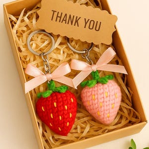 May include: Two crochet strawberry keychains, one red and one pink, with green tops and pink bows, presented in a brown gift box with a "THANK YOU" tag. The keychains are nestled in decorative straw.