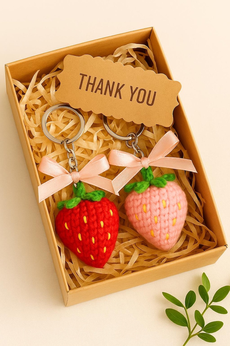 May include: Two crochet strawberry keychains, one red and one pink, with green tops and pink bows, presented in a brown gift box with a "THANK YOU" tag. The keychains are nestled in decorative straw.
