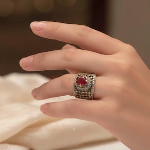 May include: An ornate ring with a large, oval red gemstone. The ring has a detailed design with small, sparkling stones and a dark metal band. The ring is displayed on a hand.