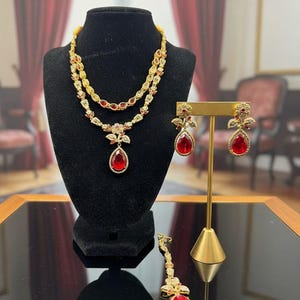 May include: A gold-toned jewelry set featuring a two-strand necklace, earrings, and a headpiece, all adorned with red teardrop-shaped gemstones and smaller clear stones. The necklace and earrings are displayed on a black velvet stand.